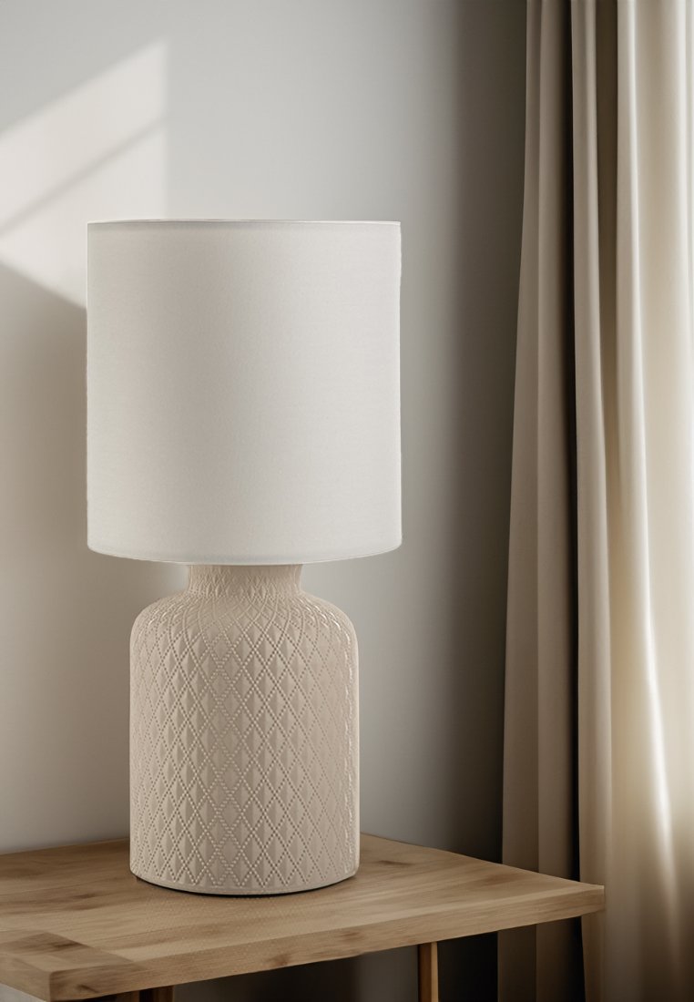 Weißer Lampenschirm aus Stoff auf einem strukturierten, beigen Keramikfuß mit Rautenmuster, platziert auf einem Holztisch in der Nähe von Vorhängen.