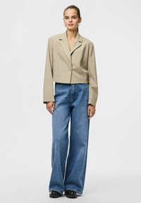 Beige croppet blazer med enkelt knaplukning, kombineret med højtaljede, bredbenede blå cowboybukser. Sorte platformssko.