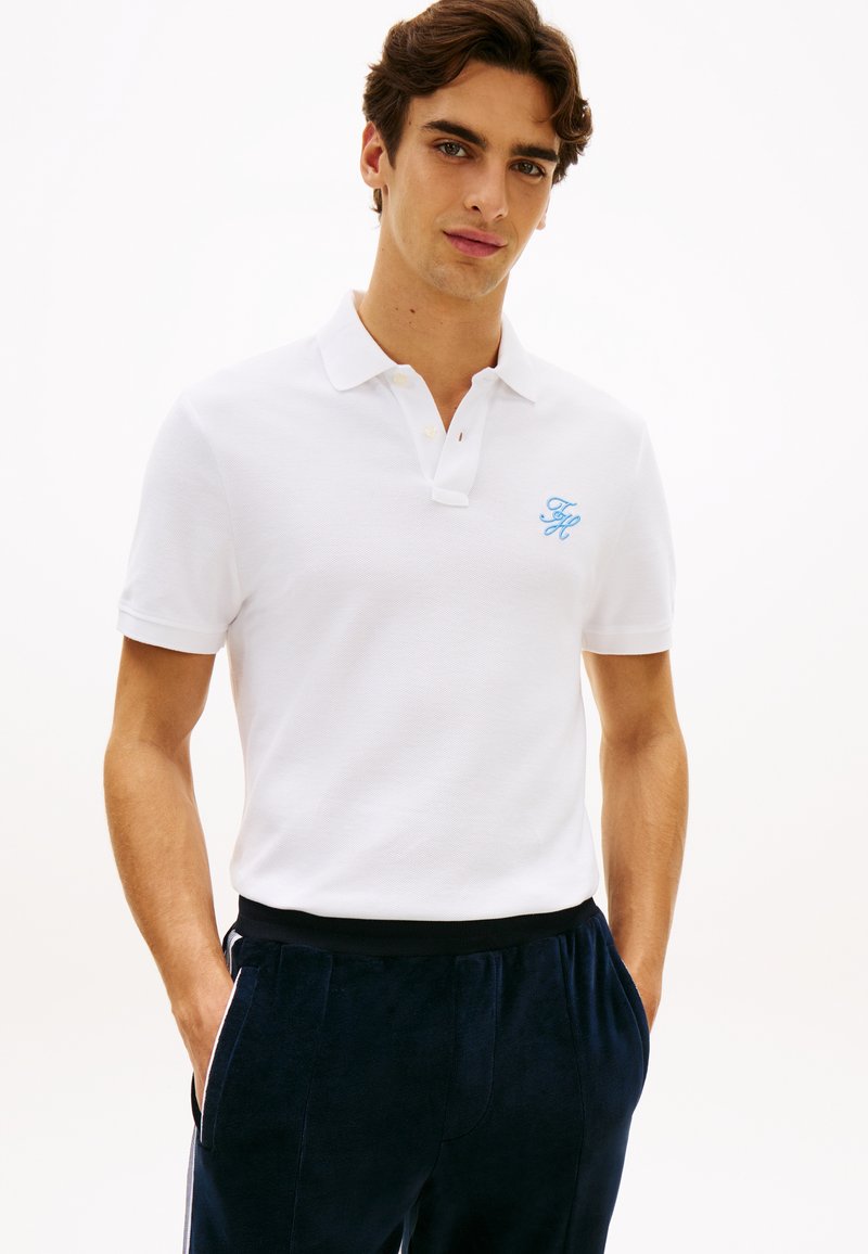 Νεαρός άνδρας με καστανά μαλλιά φοράει λευκό πόλο με ανοιχτό μπλε λογότυπο και σκούρο μπλε παντελόνι, στέκεται με τα χέρια στις τσέπες μπροστά σε λευκό φόντο.