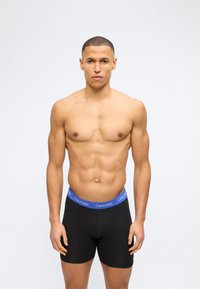 Γυμνασμένος άνδρας με κοντά μαλλιά, φορώντας μαύρα μποξεράκια με μπλε ζώνη Calvin Klein, στέκεται πρόσωπο με πρόσωπο μπροστά σε ένα απλό λευκό φόντο.