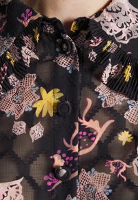 Gros plan sur un chemisier noir transparent avec des motifs floraux en jaune, rose et violet, avec des boutons recouverts de tissu et des détails de col à volants.