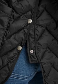 Giacca trapuntata nera con cuciture diagonali, bottoni a pressione argentati e bordi neri, abbinata a jeans in denim blu.