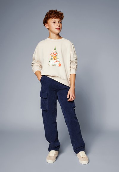 Sudadera beige con un diseño gráfico colorido. Pantalones cargo de pana en color marino con bolsillos laterales. Zapatos ligeros y texturizados.