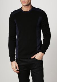 Pull noir avec des accents bleu marine sur les épaules et les côtés, col rond et poignets côtelés. Texture douce. Coupe ajustée.