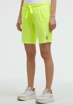 Person trägt neongelbe Shorts mit Kordelzug und Taschen sowie weiße beige Sneakers und steht vor einem einfarbigen Hintergrund.
