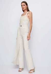 Tuta bianca monospalla con design drappeggiato, corpetto aderente e gambe svasate. Presenta un dettaglio metallico in vita e una texture setosa.