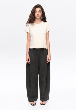 Mujer con cabello largo y rizado que lleva una camiseta blanca de manga corta, pantalones sueltos anchos de color gris oscuro y chanclas negras, de pie contra un fondo blanco.