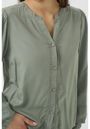 Bluzka z długim rękawem w kolorze szałwi zielonej z obszytymi tkaniną guzikami i koronkową lamówką wzdłuż listwy guzikowej i mankietów, noszona przez osobę.