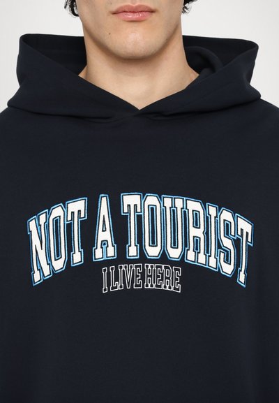 Fekete kapucnis pulóver elülső grafikával, amelyen a "NEM TURISTA" és az "ITTHON LAKOM" feliratok fehér és világoskék betűkkel olvashatók. Puha pamut anyagból készült, laza fazonú.