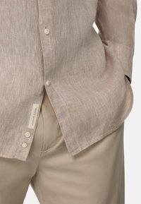 Camicia in lino beige chiaro con finitura testurizzata, dotata di colletto classico, pattina con bottoni e piccolo etichetta con la scritta "Lino Francese".