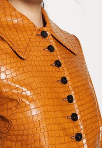 Primo piano di una persona che indossa una giacca di pelle color tan con una texture a motivo coccodrillo e bottoni neri allacciati sul davanti.
