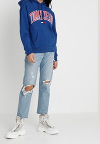 Sudadera azul con el texto "TOMMY JEANS" en rojo, bolsillo, combinada con jeans claros desgastados que presentan desgarros en las rodillas y zapatillas altas blancas.