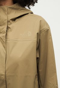 Persona che indossa una giacca impermeabile beige di The North Face con cappuccio e logo del marchio sul petto.