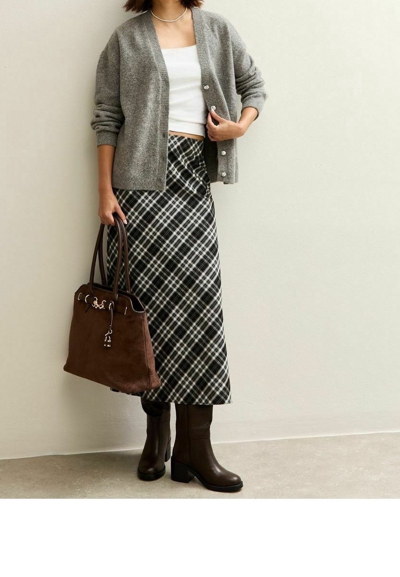 Femme portant un cardigan gris sur un haut blanc, une jupe à carreaux, des bottes marron foncé, tenant un grand sac à main marron contre un mur uni.