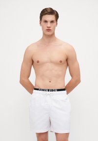 Bijele kupaće hlače od laganog materijala, s crnim elastičnim pojasom na kojem je logo "CALVIN KLEIN". Ravnog kroja, bez uzoraka.
