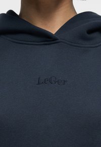 Κοντινή λήψη μπλε ναυτικού φούτερ με ανάγλυφο κεντημένο κείμενο "LeGer" στο στήθος, που φοριέται από άτομο που φαίνεται από τον λαιμό έως τον άνω κορμό.