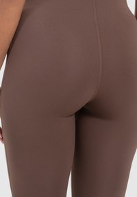 Bruine leggings van rekbaar materiaal, met een gladde textuur en contrasterende stiksels aan de zijkanten en de tailleband.