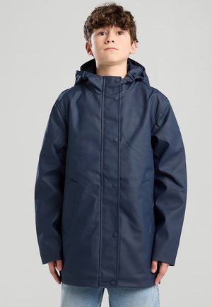 Jongen met krullend bruin haar, gekleed in een marineblauwe regenjas met capuchon, knopen aan de voorkant en zakken, staand tegen een effen achtergrond.