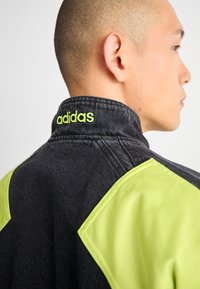 Jaqueta de ganga preta com detalhes em verde neon nos ombros e logótipo no colarinho. Apresenta um colarinho alto e um contraste de cores distinto entre os materiais.