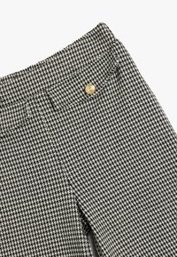 Hahnentritt-Muster in Schwarz und Weiß auf einem Stoff, mit Nahaufnahme, die die Textur und ein goldenes Knopfdetail am Bund zeigt.