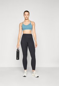 Sportos öltözet, amely magában foglal egy világoskék sportmelltartót, fekete, magas derekú leggings-t és fehér tornacipőt; a kezében egy fekete vizespalackot tart.