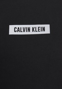 Tissu noir à côtes présentant une étiquette rectangulaire blanche avec le texte "CALVIN KLEIN" en lettres noires en gras.