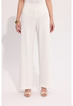 Kobieta ubrana w białe, spodnie z wysokim stanem i szerokimi nogawkami oraz beżowe sandały na obcasie, z złotą bransoletką na lewej ręce, stojąca na białym tle.