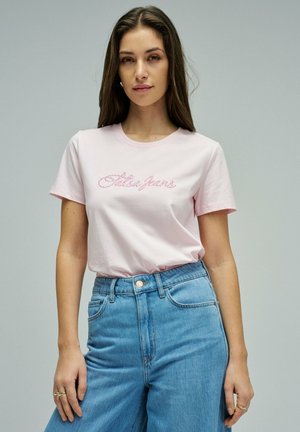 Mujer con el pelo largo y castaño que lleva una camiseta rosa claro "Salsa Jeans" metida dentro de unos vaqueros azules de talle alto, de pie frente a un fondo liso.