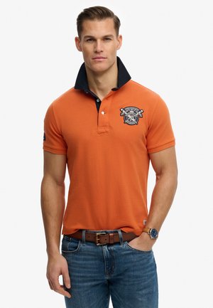 Mężczyzna w pomarańczowej koszulce polo z podniesionym czarnym kołnierzem, niebieskich dżinsach, brązowym pasku i zegarku na nadgarstku na białym tle.