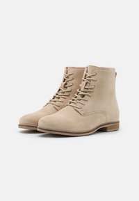 Bottines en daim beige avec bout arrondi, devant à lacets et petit talon en bois. Texture lisse et design minimaliste.