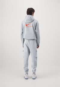 Šedá mikina s kapucí a červeným logem Nike, doplněná o odpovídající tepláky s kapsami a bílým logem. Nosit s obuví světlé barvy.
