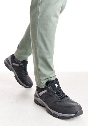 Schwarze Sportschuhe mit grauen Akzenten, featuring ein strukturiertes Obermaterial, gepolsterte Sohle und Schnürdesign, kombiniert mit hellgrünen Hosen.