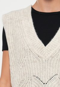 Jasno beżowy sweter bez rękawów z dekoltem w kształcie litery V, z teksturowanym wzorem i szerokimi ramiączkami, warstwowy na czarnym topie z prążkowanego materiału.