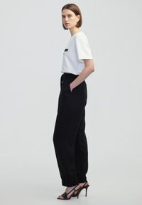 Witte overhemd met korte mouwen en zwarte tekst, gecombineerd met losse zwarte broek met een elastische tailleband, en zwarte hoge hakken.