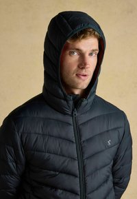 Mężczyzna o jasnych włosach i niebieskich oczach, ubrany w czarną pikowaną kurtkę z kapturem, lekko patrzący w bok na tle jednolitego beżowego tła.