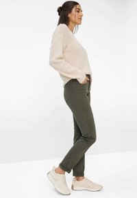 Beiger, gestrickter Pullover, olivgrüne, schmal geschnittene Hose und helle Sneaker mit Gummisohlen. Seitenansicht vor einem schlichten weißen Hintergrund.