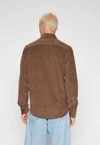 Mies, jolla on lyhyet vaaleat hiukset, pukeutuneena ruskeaan samettipaita long-sleeve ja vaaleansinisiin farkkuihin, nähtynä takaa yksinkertaista taustaa vasten.