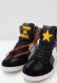 Vysoké tenisky s černým koženým tělem, semišovými akcenty a bílou gumovou podrážkou. Zdůrazněný žlutý nápis "TOHLE JE SKUTEČNÉ" a hvězdička. Bordó tkaničky.