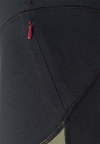 Detailní záběr na černou sportovní látku s propracovaným švem a malým červeným táhlem na zipu na kapse.