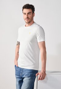 T-shirt blanc à manches courtes avec un col rond et un petit emblème doré sur la poitrine, associé à un jean en denim bleu. Tissu lisse, design simple.