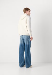 Hoodie in hellbeige, kombiniert mit lässigen, weit geschnittenen blauen Jeans mit einem verwaschenen Design und Distressed-Details. Bequeme Schuhe runden den Look ab.