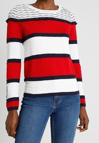 Pull rayé rouge et blanc avec un motif texturé, accents bleu marine, et un col côtelé ; présente des volants aux épaules pour plus de détails.