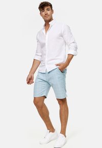 Witte linnen blouse met knopen, lichtblauwe geruite shorts en witte sneakers. De kleding is casual met een comfortabele pasvorm.