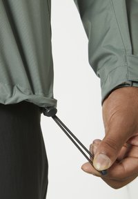 Chaqueta de tela verde claro con un cordón ajustable en el dobladillo. Una mano sujeta un tope negro, ajustando el cordón elástico para un mejor ajuste.