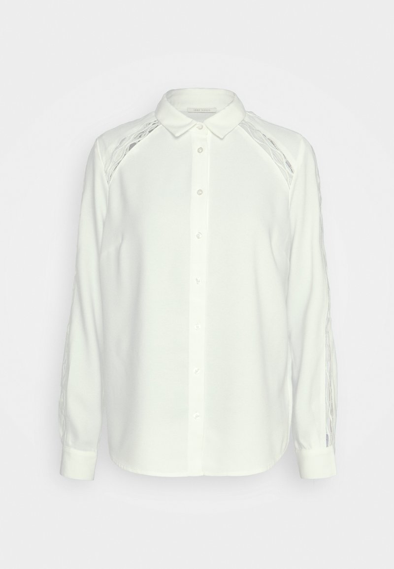 Blouse blanche à manches longues boutonnée avec col, avec des détails en dentelle ajourée le long des épaules et des manches extérieures.