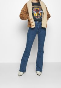 Brun mockasinsjacka med krämfärgad fuskpälskrage, svart grafisk t-shirt, högmidjade blå utsvängda jeans och vita spetsiga ankelstövlar.