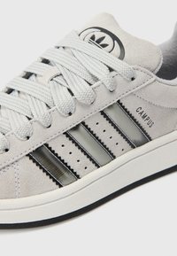 Szürke velúr sneaker, három fekete csíkkal, texturált részletekkel és dombornyomott logóval a nyelvén. Fehér középtalpas, fekete külsőtalpas.