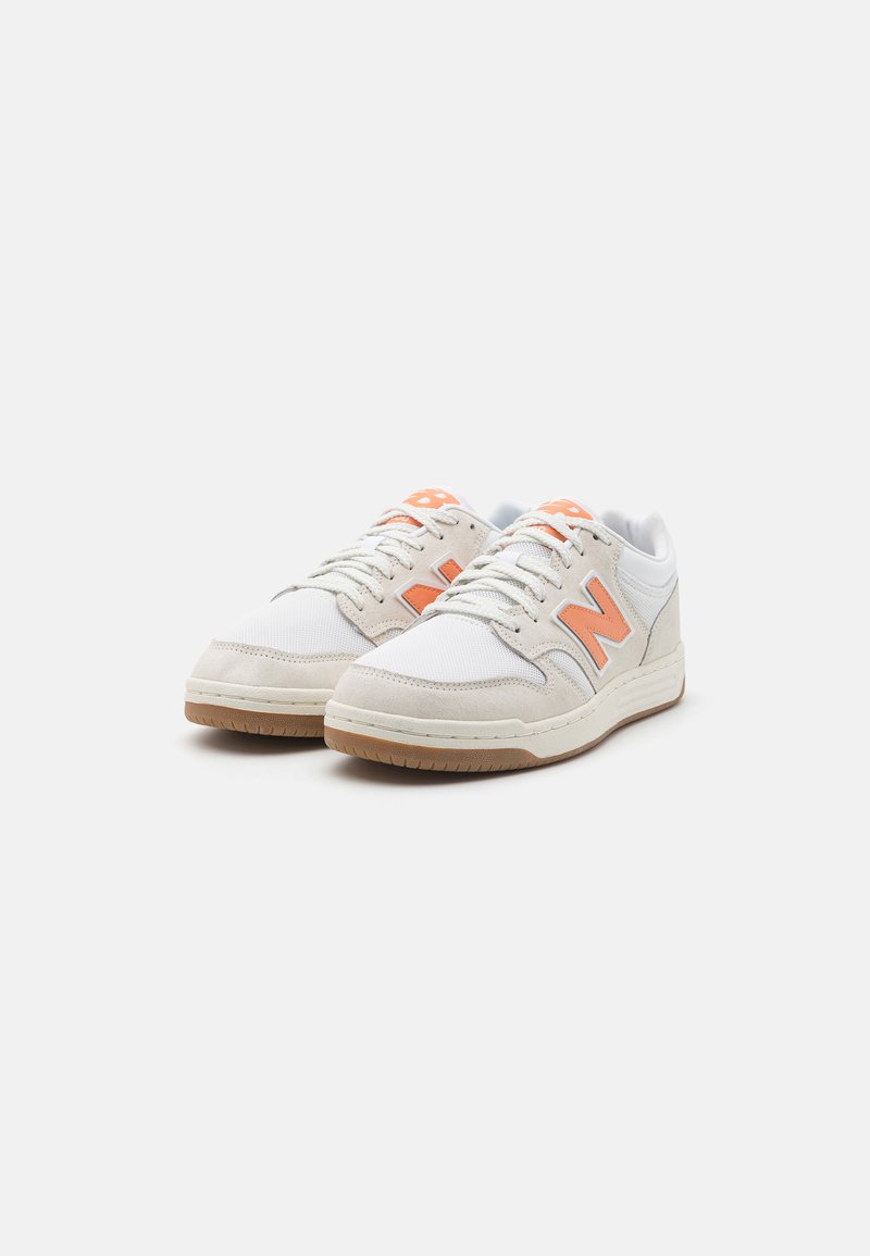 Baskets sportives blanches et beiges avec des accents orange, panneaux en suède texturé et en maille, semelle en caoutchouc et un logo "N" bien en vue sur le côté.