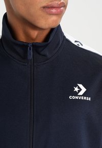 Navy blå zip-jacka med hög krage, som har vita detaljer på axlarna och en logotyp med en stjärna och pil. Slät tygstruktur.