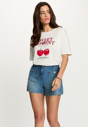 Kvinde med langt mørkt hår iført en hvid t-shirt med røde kirsebær og tekst, kombineret med blå denimshorts og guldsmykker på hånden.